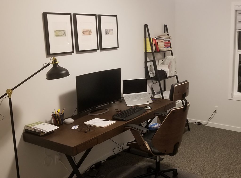 A wooden desk in a home office with chair, floor lamp, and shelves.