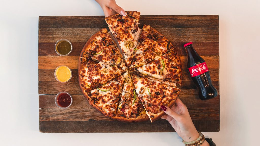 two hands pulling slices of pizza off a cutting boards with 3 dipping sauces and a bottle of coca cola